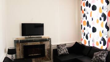 A living room with a flat-screen TV mounted above a tiled fireplace, a black sofa with patterned cushions, and a colorful curtain.