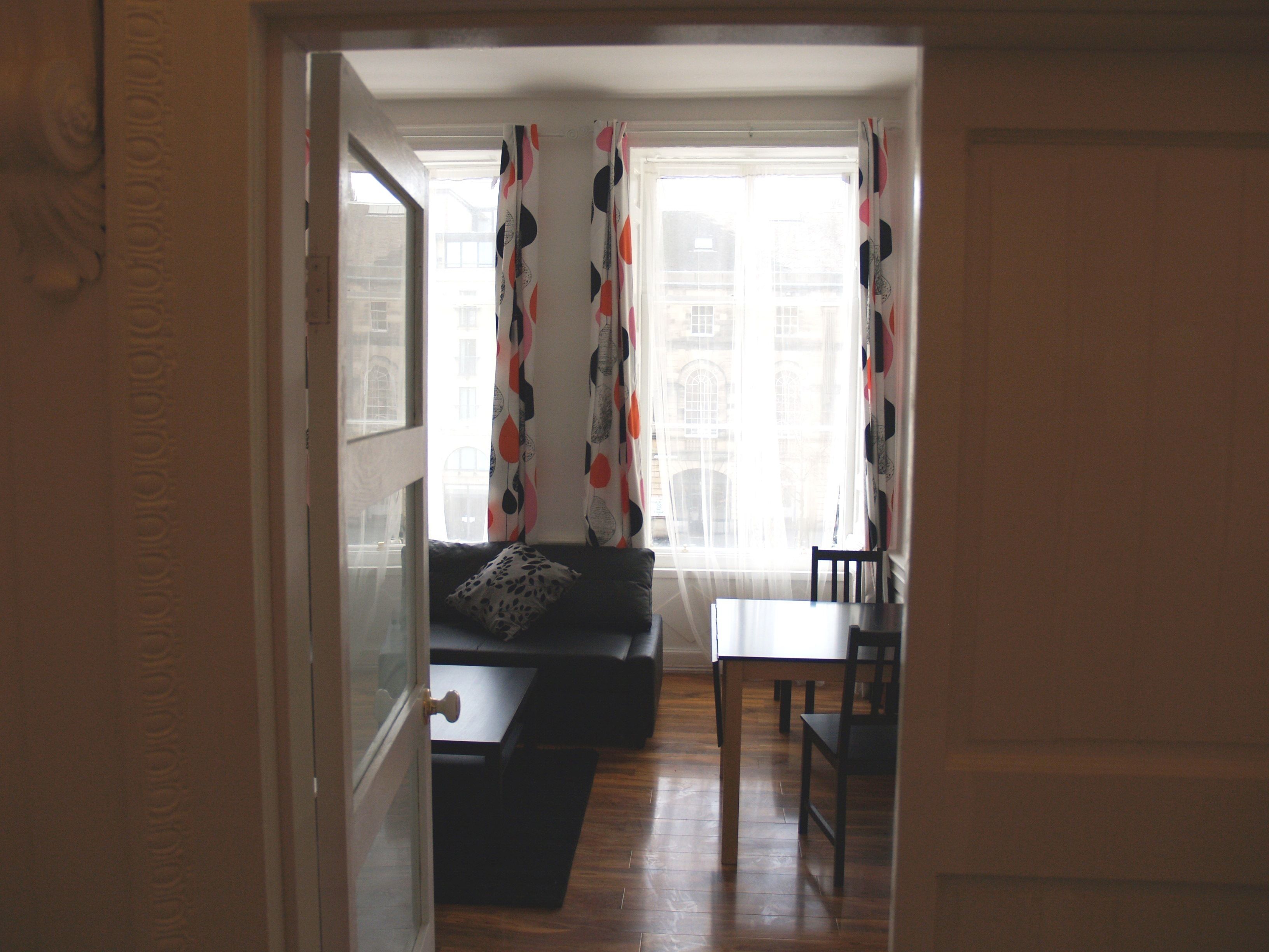 Una habitación con piso de madera, un sofá, una mesa y un ventanal con cortinas estampadas.