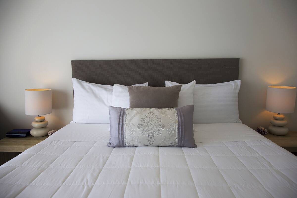Una cama bien hecha con edredón blanco, una almohada decorativa y dos lámparas de noche sobre mesitas de madera.