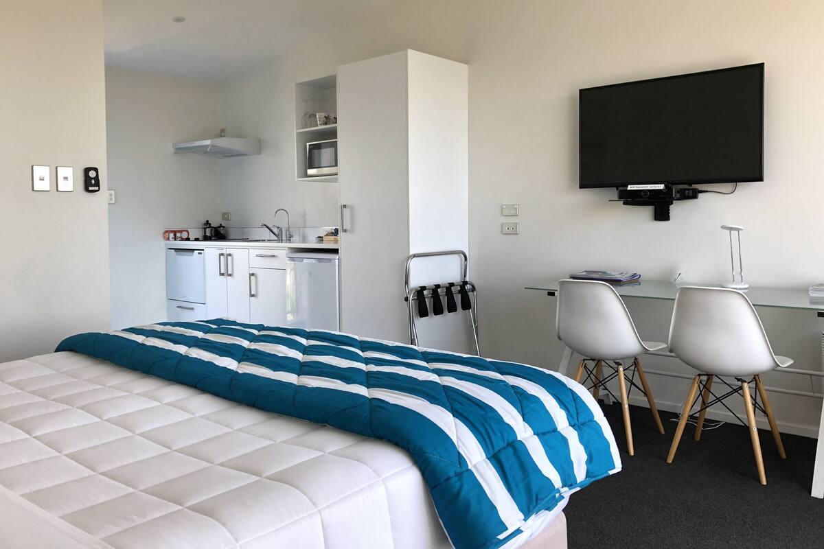 Una habitación de hotel moderna con cama, escritorio, dos sillas, televisor y kitchenette.