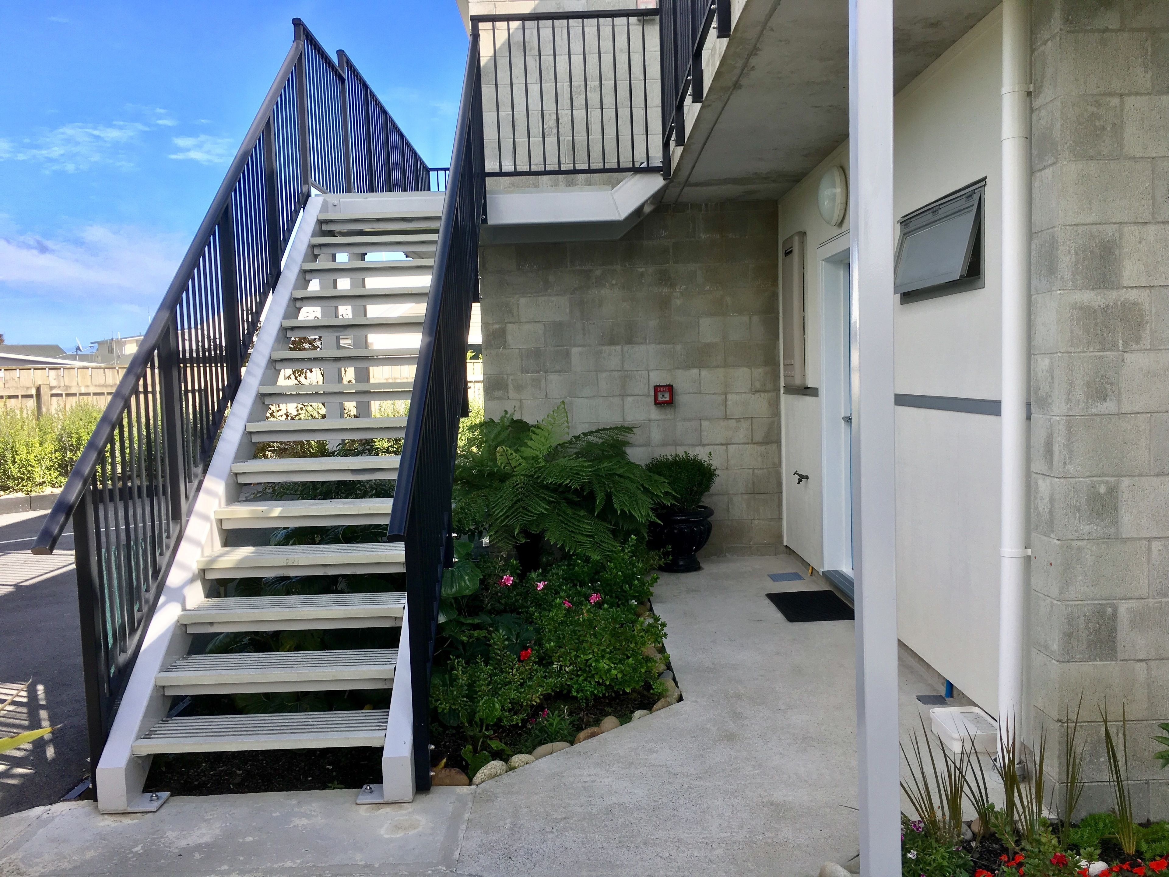 A staircase leading to a building with a fire alarm on the wall, a door, and a small garden area with plants and flowers.