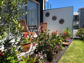 Un patio con sillas y mesa de madera, rodeado de plantas y flores en macetas.