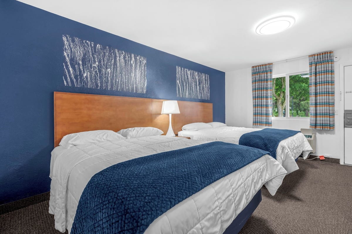 Une chambre d’hôtel avec deux lits, une tête de lit en bois, une lampe blanche et une vue sur la verdure par la fenêtre.