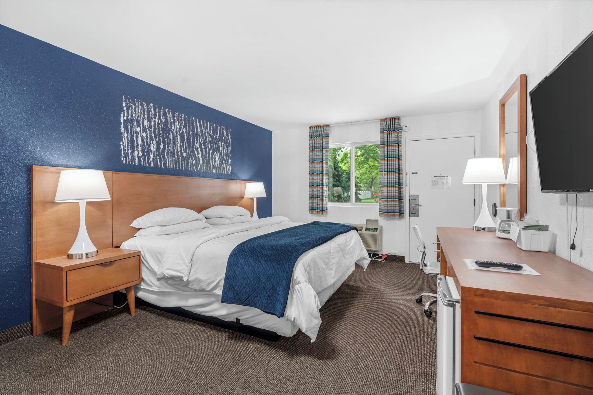 Una habitación de hotel con cama, mesita de noche, lámpara, cómoda, espejo, televisor y ventana con cortinas.