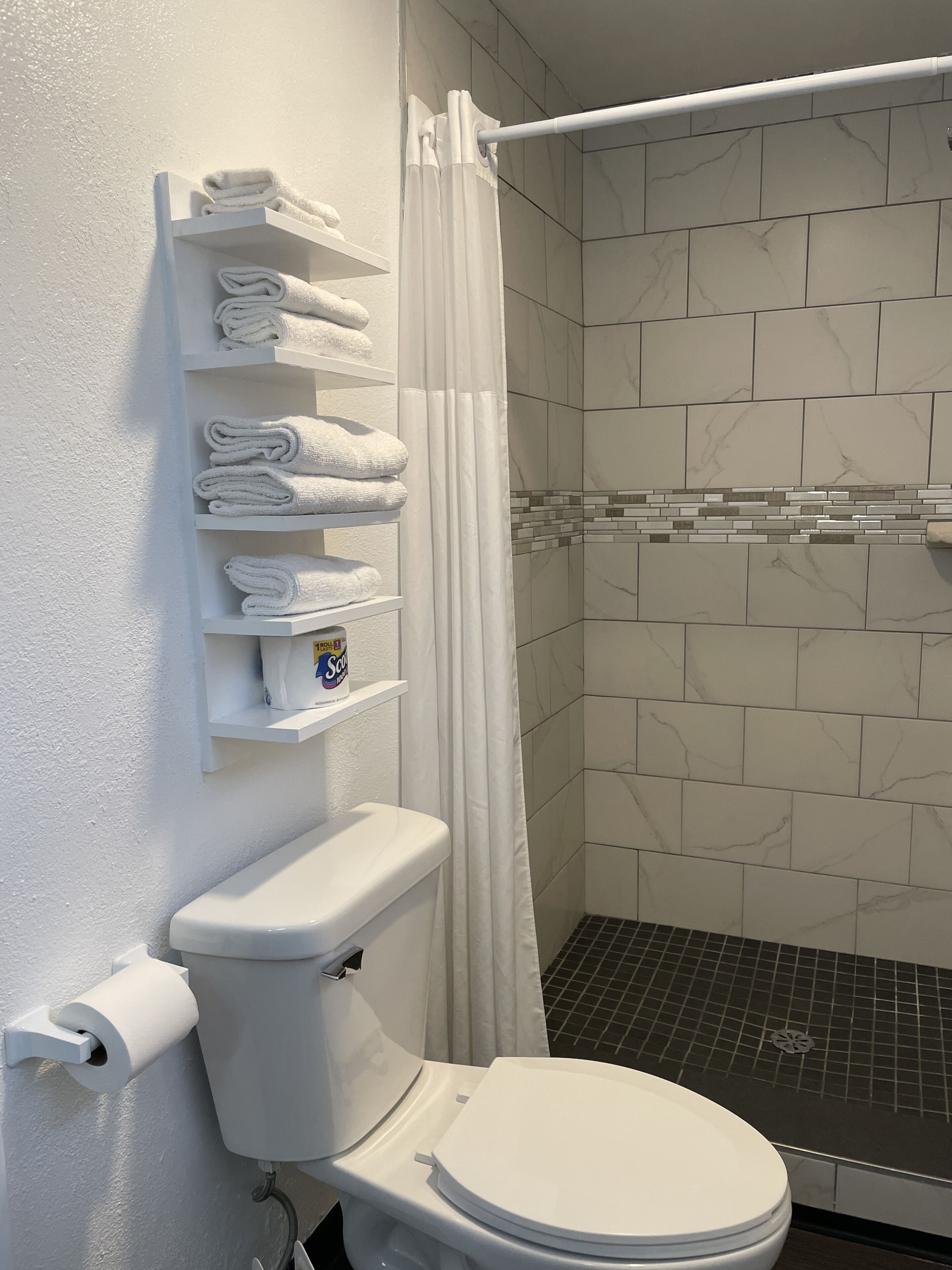 A bathroom with a toilet, a shelf with folded towels, a shower curtain, and a tiled shower area.