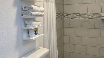 A bathroom with a toilet, a shelf with folded towels, a shower curtain, and a tiled shower area.