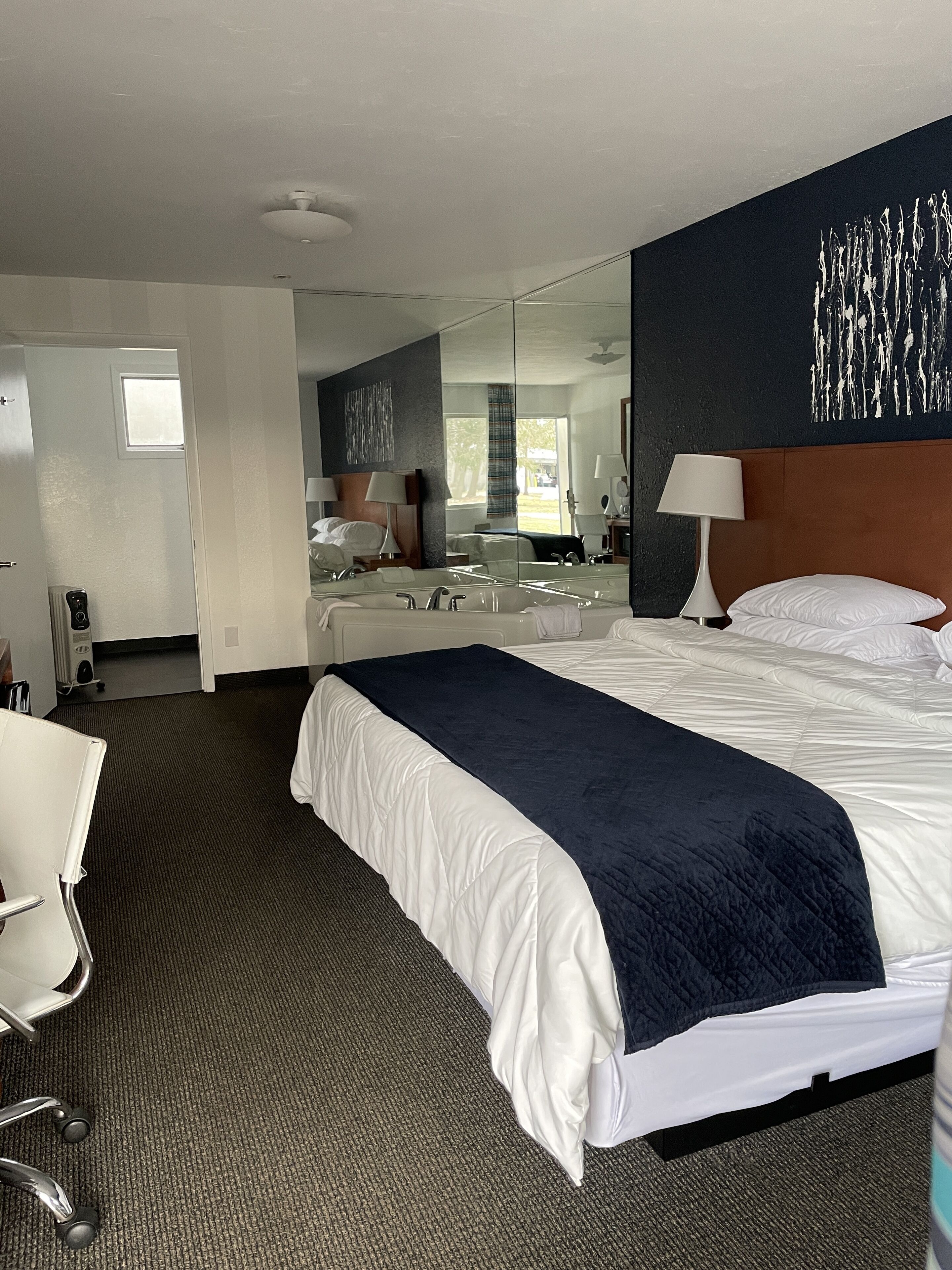 Une chambre d’hôtel avec un grand lit, un bureau et une chaise. Il y a une fenêtre donnant sur des arbres et une porte qui mène à une autre pièce.
