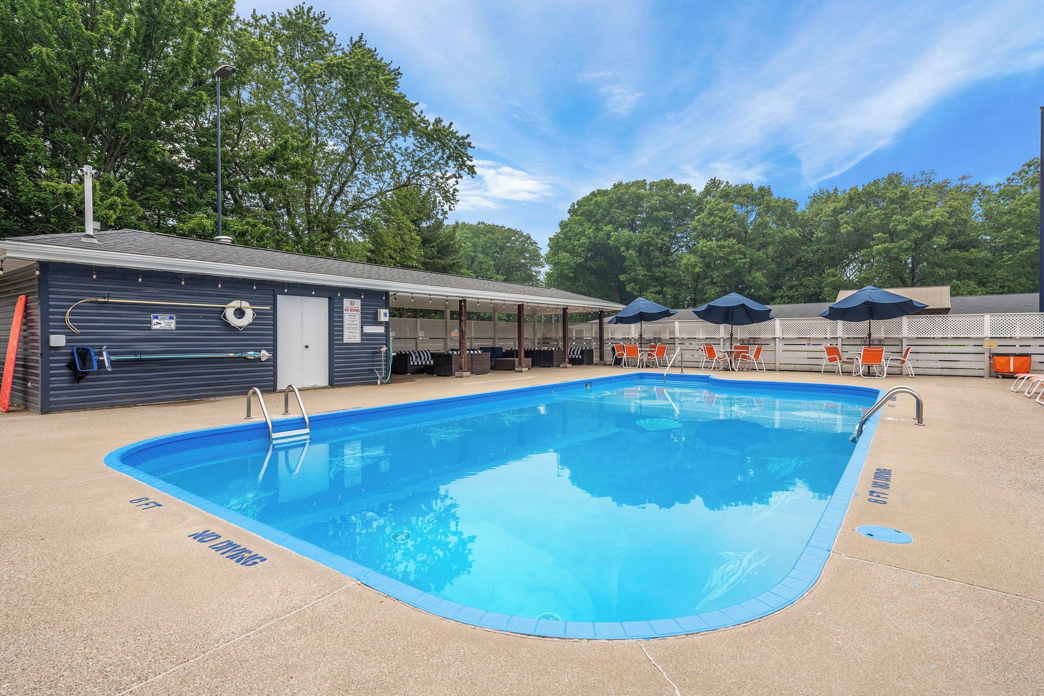 A swimming pool with a covered area and lounge chairs, surrounded by trees and a building with a sign.
