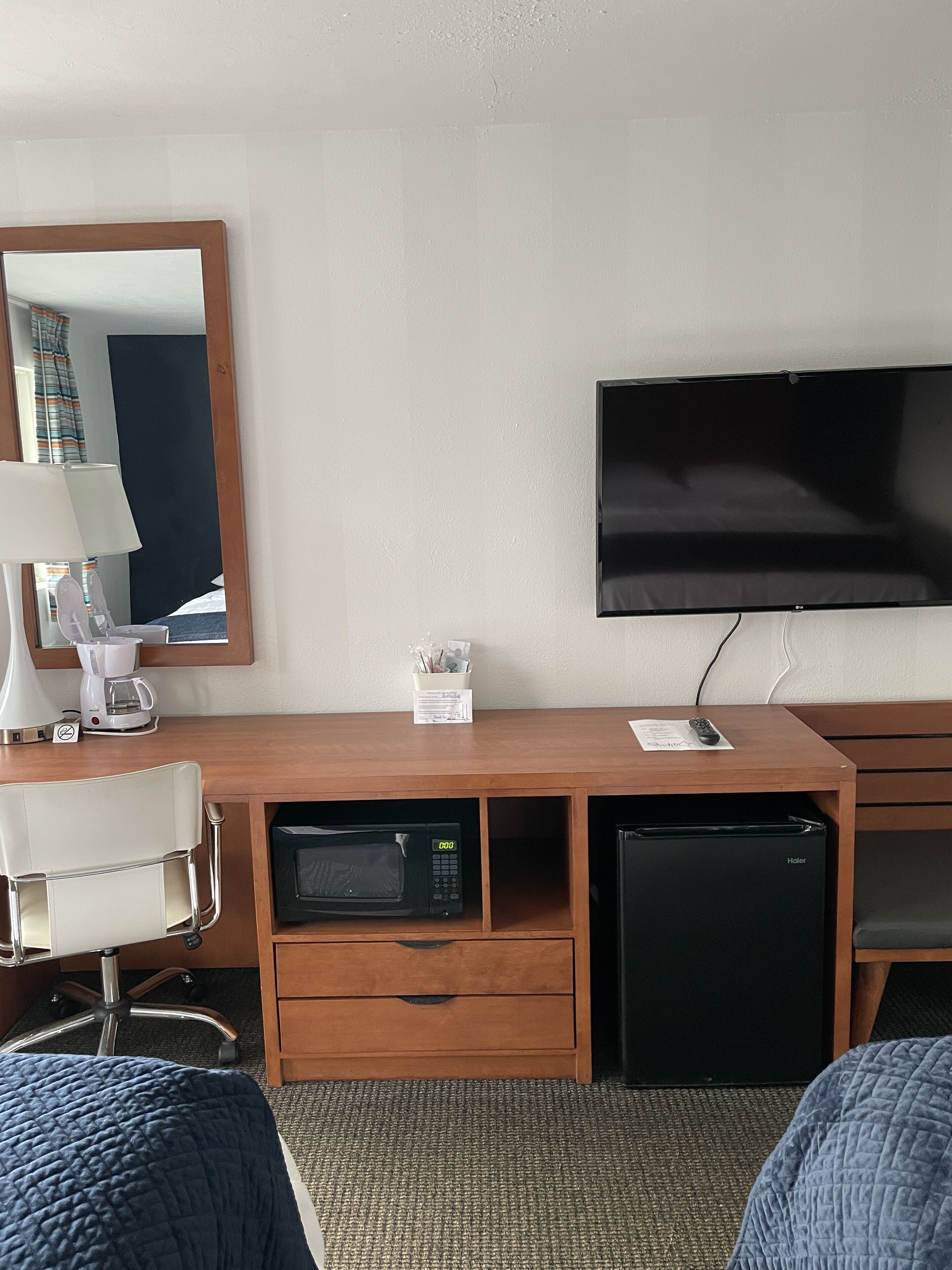 A hotel room with a wooden desk, a flat-screen TV, a microwave, a mini-fridge, a coffee maker, a lamp, and a chair.