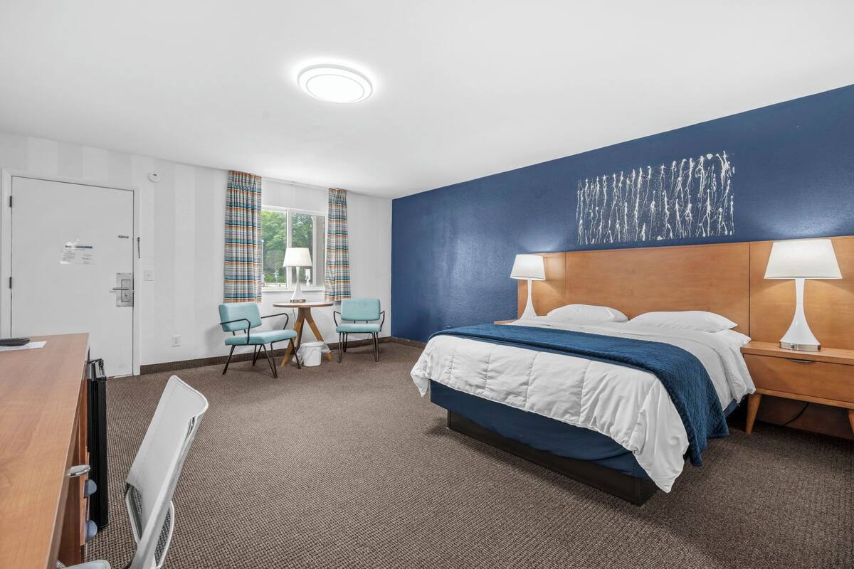 Une chambre d’hôtel avec un lit, un bureau, deux chaises, une petite table et une fenêtre avec des rideaux.