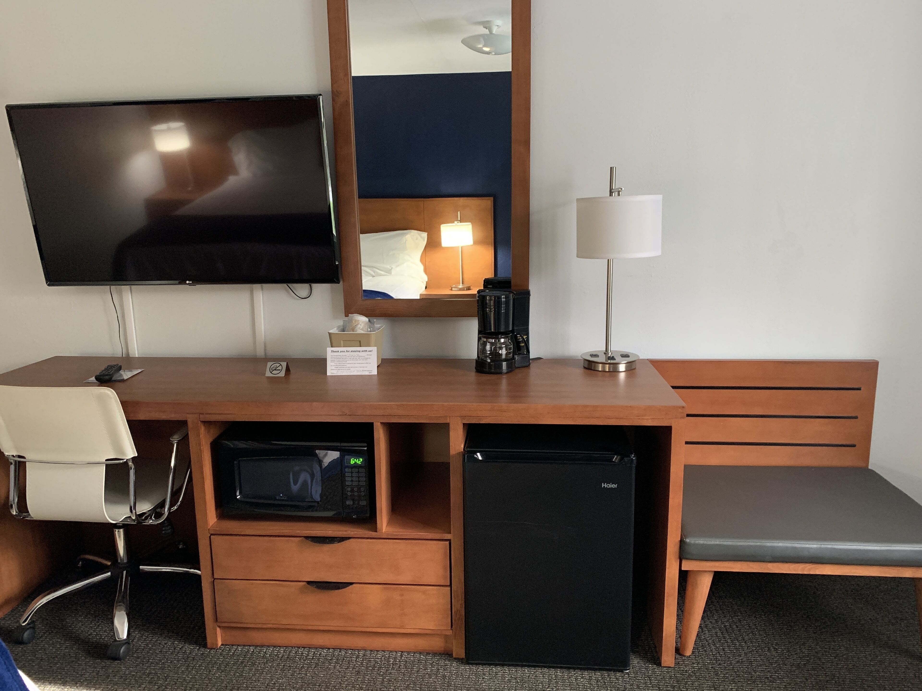 A hotel room with a wooden desk, a television, a microwave, a coffee maker, a lamp, a chair, and a small refrigerator.