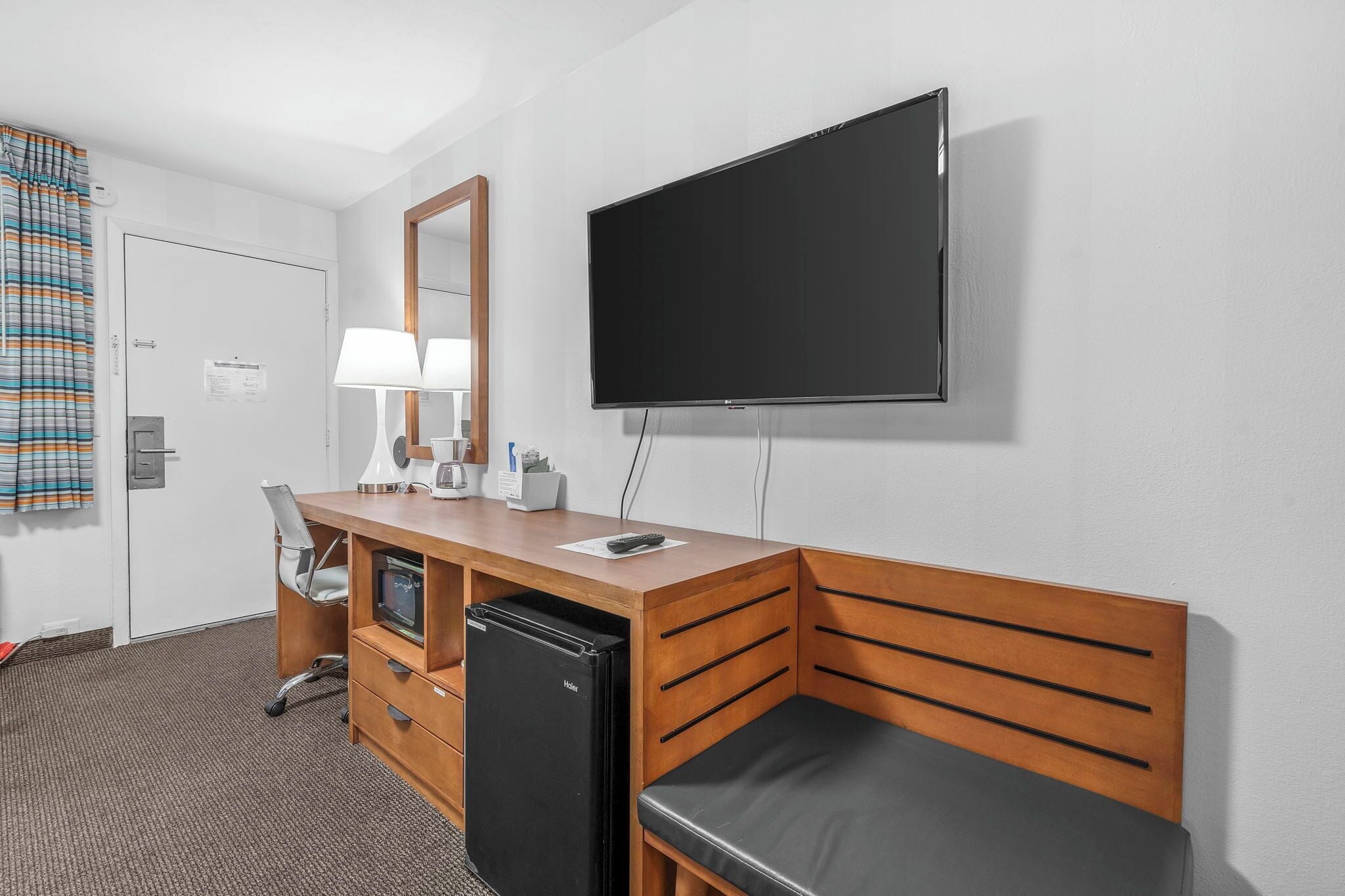 A hotel room with a wooden desk, a flat-screen TV, a mini-fridge, a chair, a bed, and a window with curtains.