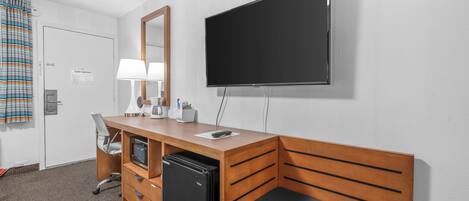 A hotel room with a wooden desk, a flat-screen TV, a mini-fridge, a chair, a bed, and a window with curtains.