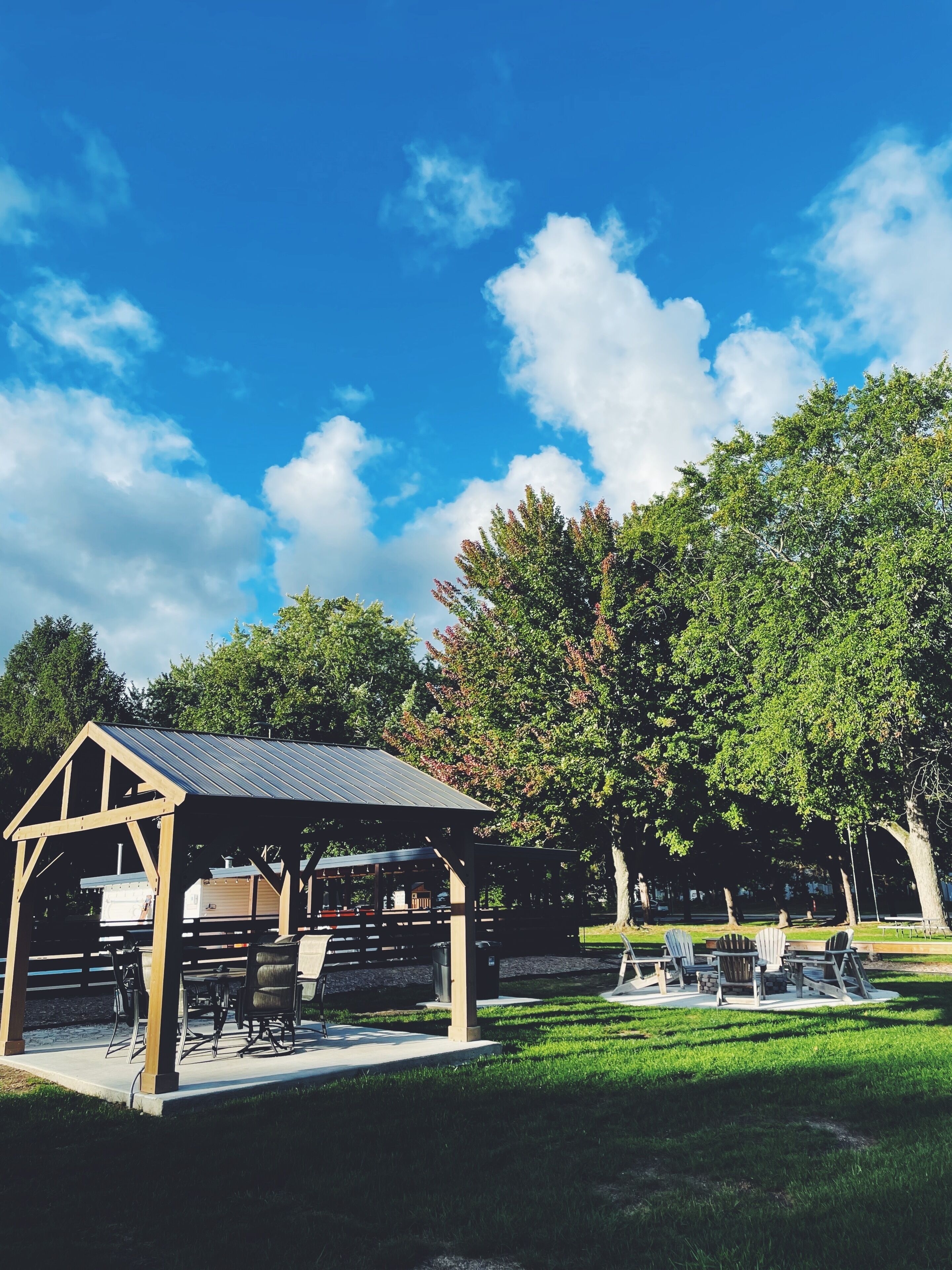 A covered picnic area with tables and chairs, surrounded by trees and a clear blue sky.