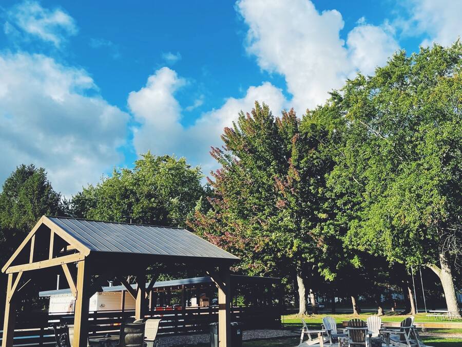 A covered picnic area with tables and chairs, surrounded by trees and a clear blue sky.