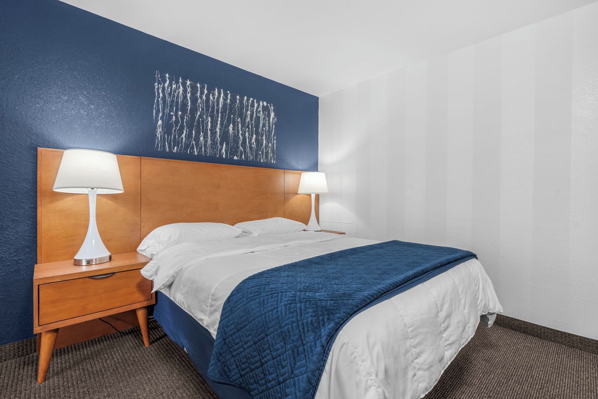 A hotel room with a wooden headboard, two bedside lamps, a bed with white and blue bedding, and a blue wall with a white abstract painting.