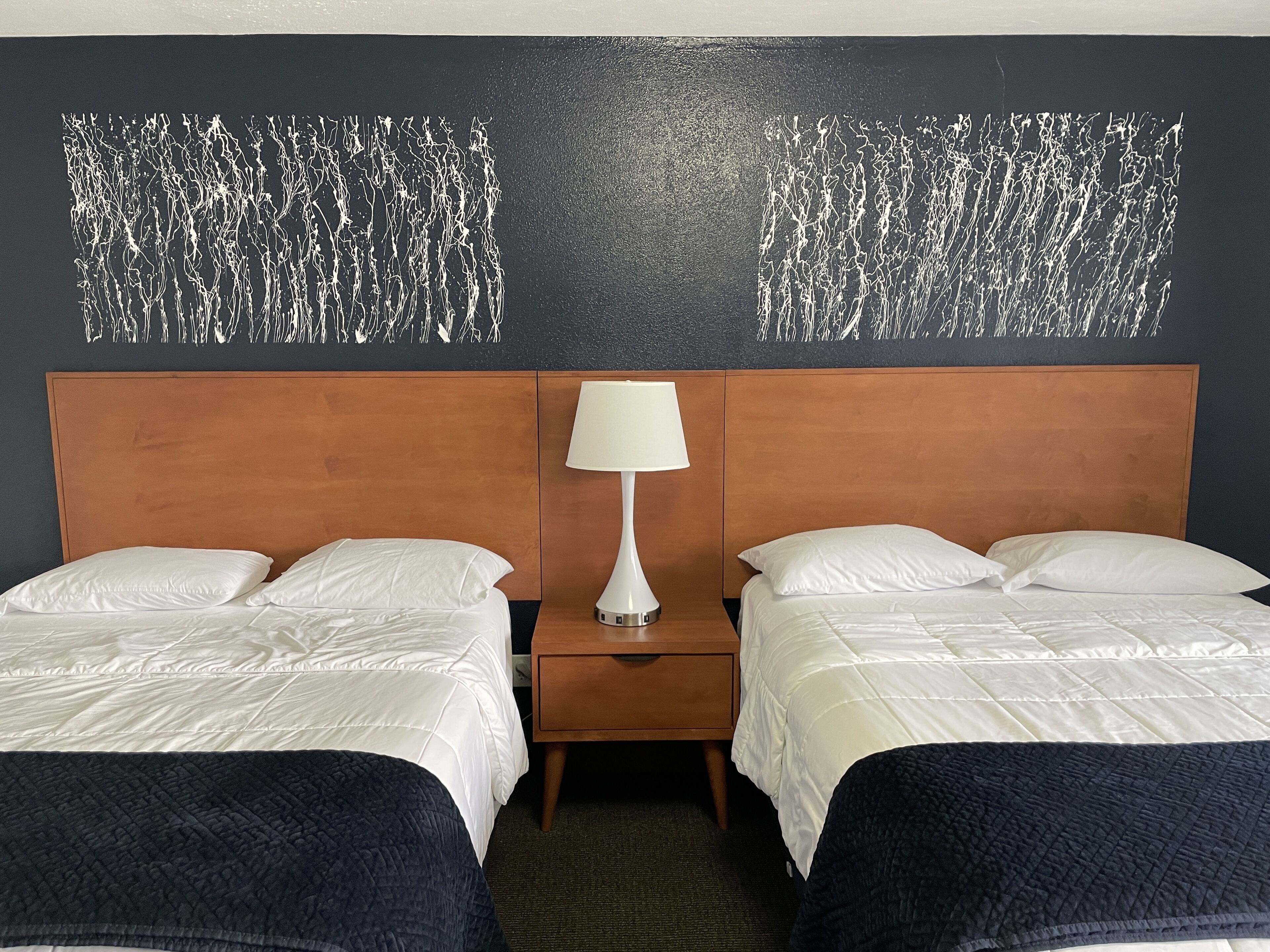 A hotel room with two beds, a wooden headboard, a nightstand with a lamp, and a textured wall decoration.