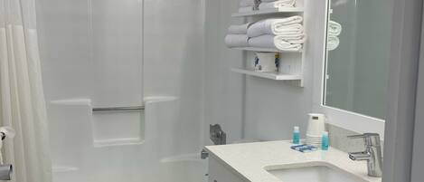 A modern bathroom with a white bathtub, a toilet, and a sink with a mirror. There are shelves with towels and toiletries.