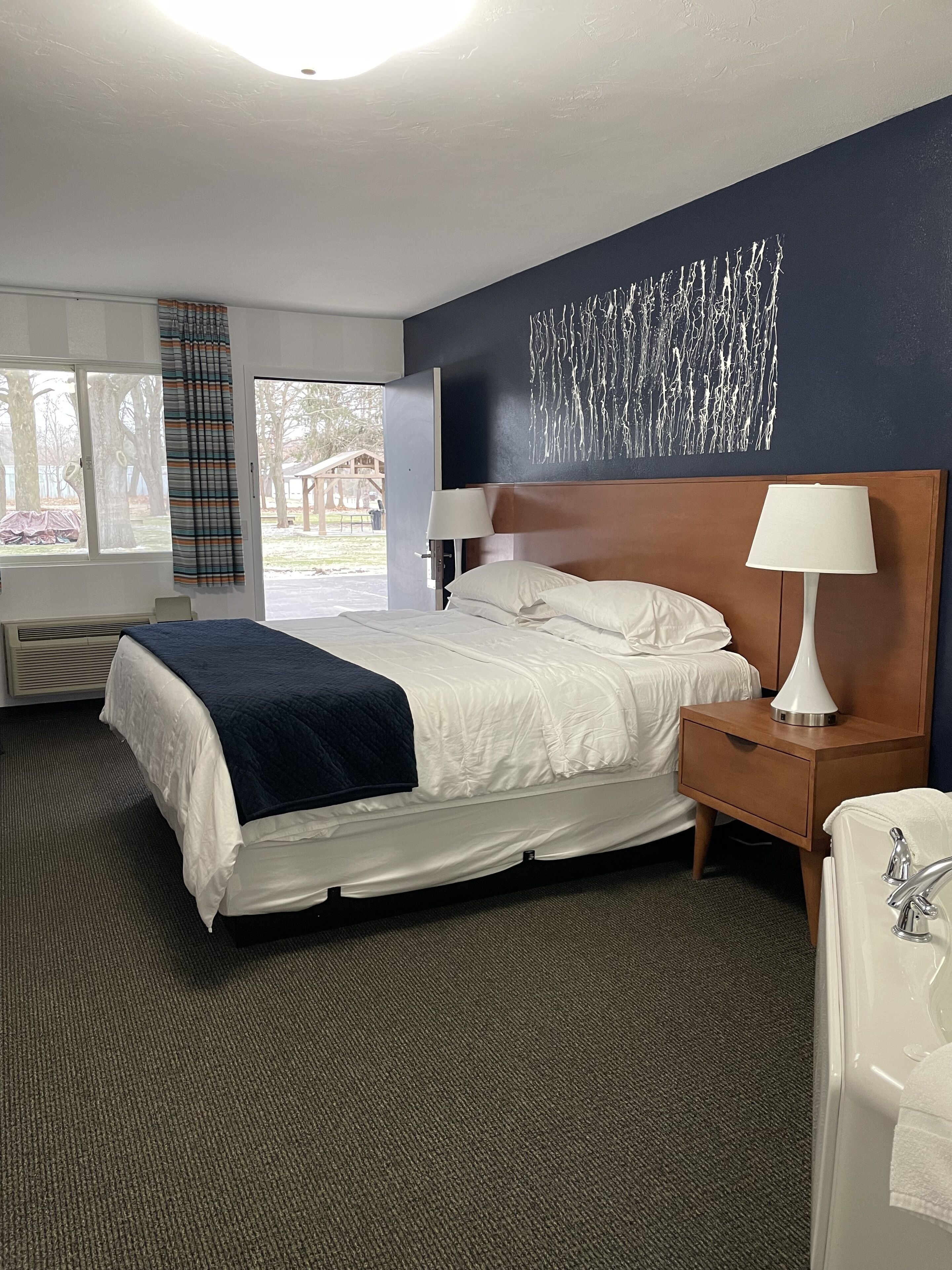Une chambre d’hôtel comprenant un lit, une table de chevet avec une lampe, une salle de bain avec un lavabo et un miroir, et une vue sur l’extérieur par une fenêtre.