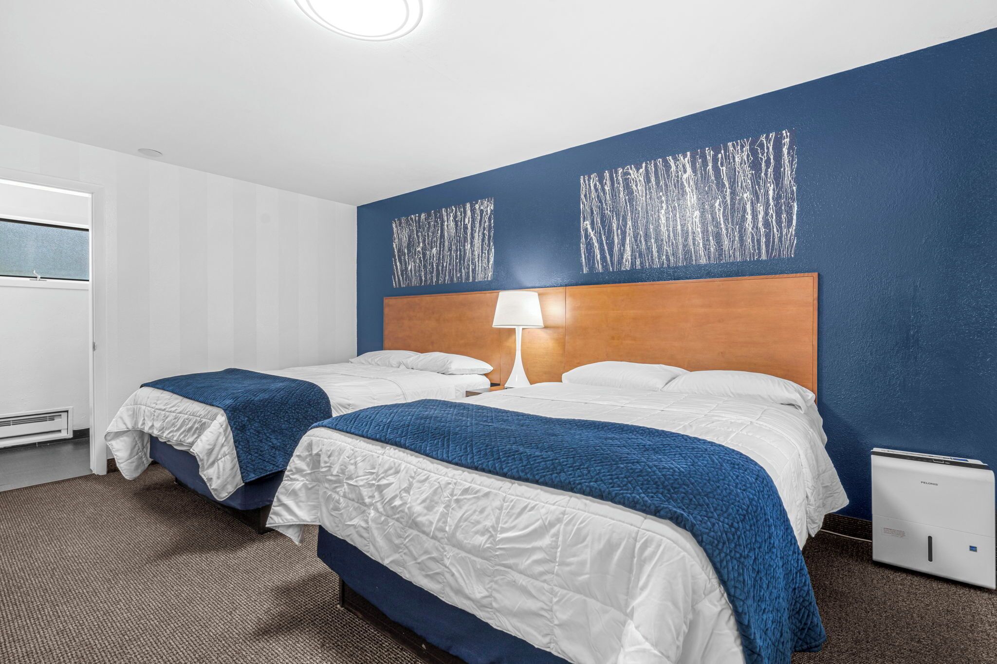 A hotel room with two beds, a wooden headboard, a white lamp, and a small bedside table.