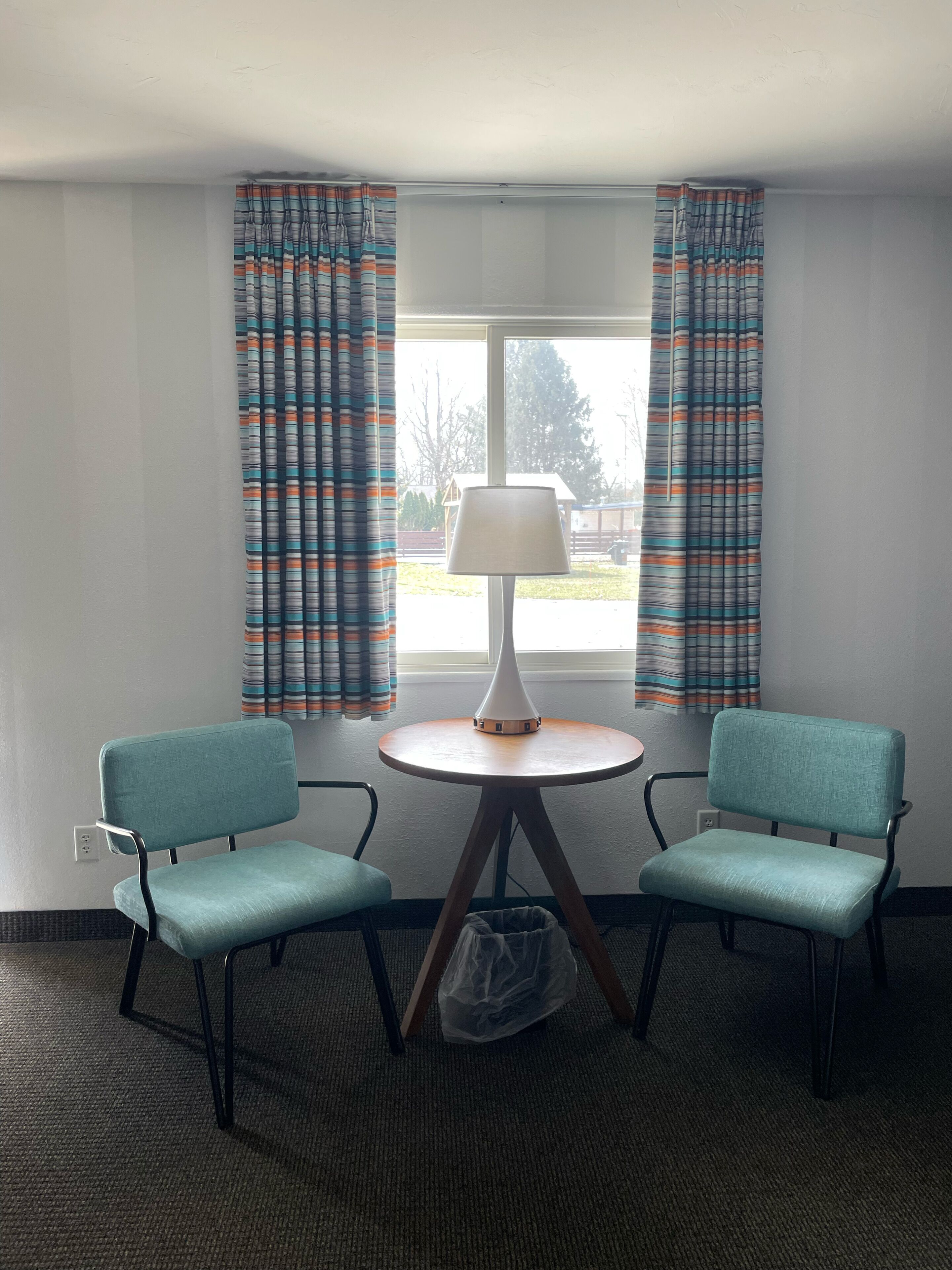 A hotel room with two teal chairs, a small round table with a lamp, and a window with striped curtains.