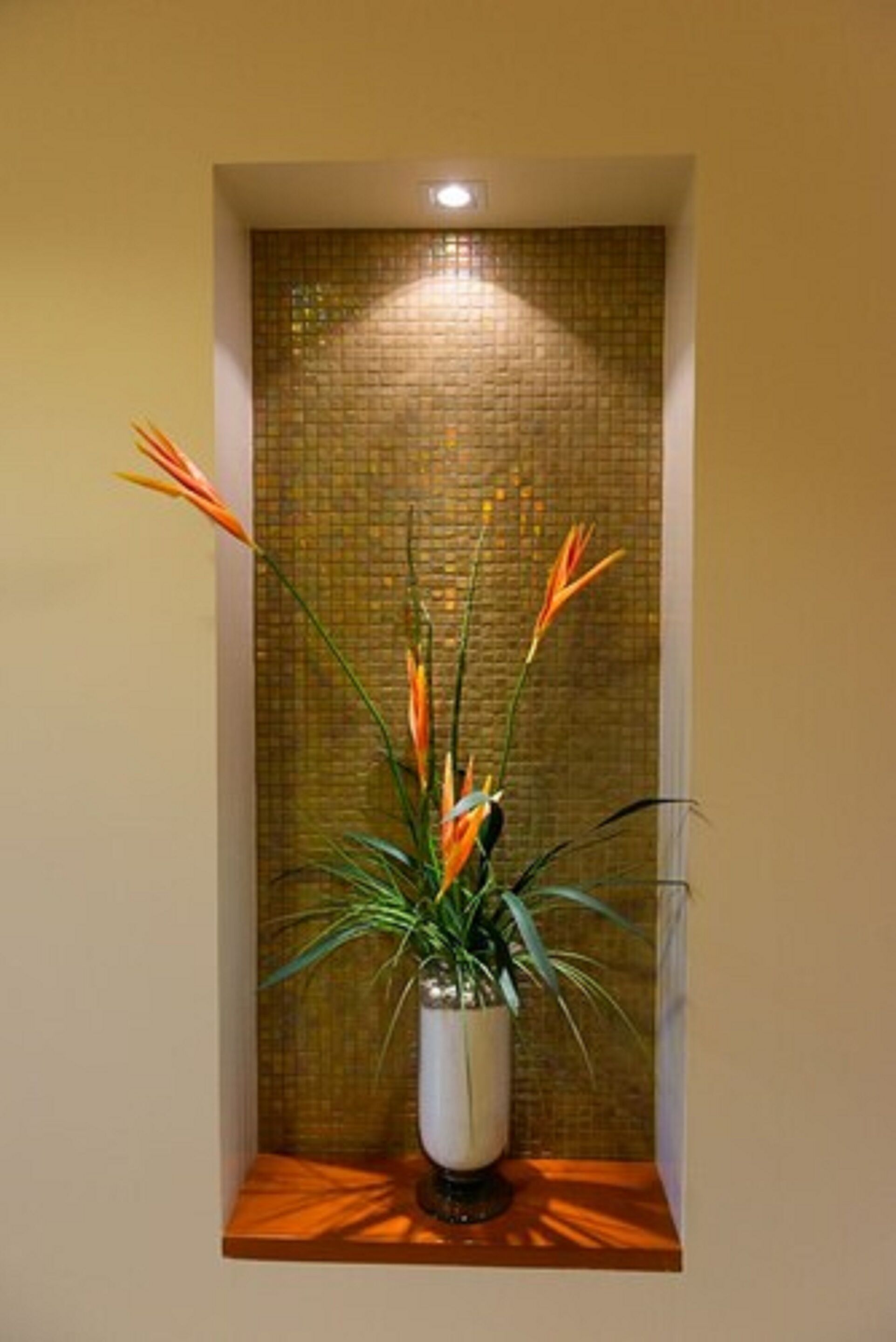 A potted plant with orange flowers in a white vase on a wooden shelf against a textured wall.