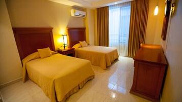 Habitación de hotel con dos camas, cabecera de madera, mesita de noche, lámpara, unidad de aire acondicionado y ventana con cortinas.