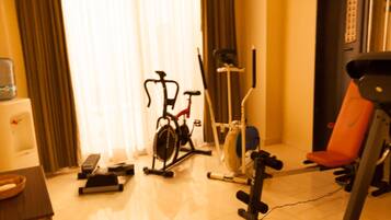 A room with a treadmill, stationary bike, and exercise equipment. There is a window with curtains, a table with a bowl, and a chair.