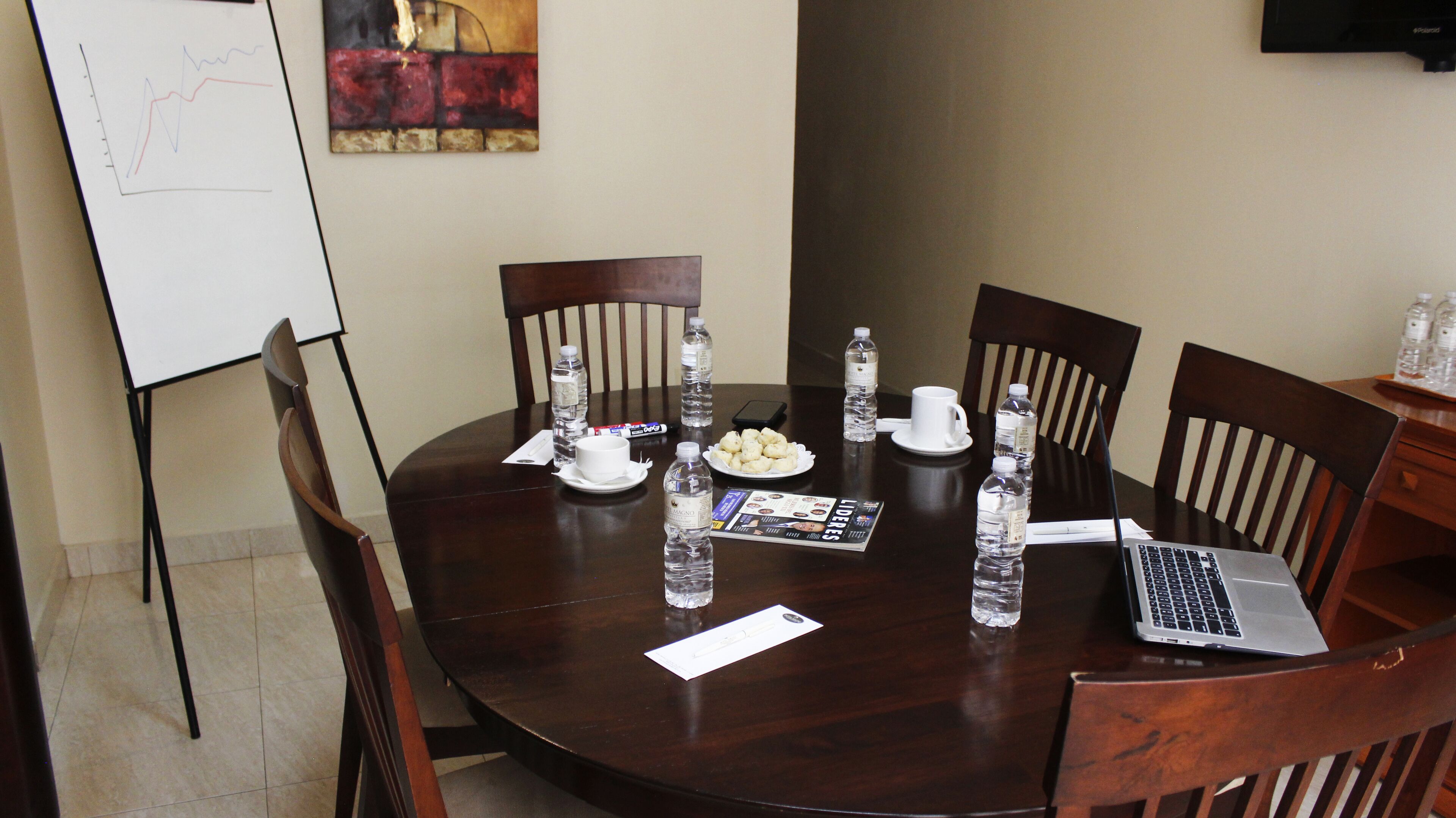 Un salón de conferencias con una mesa redonda, sillas, una computadora portátil, botellas de agua, un plato de refrigerios, una pizarra y un cuadro en la pared.