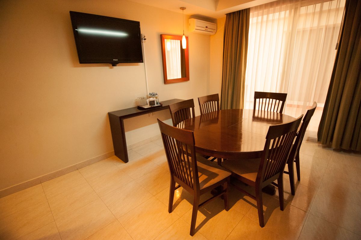 A dining area with a round table, six wooden chairs, a flat-screen TV mounted on the wall, a small table with a lamp, and a window with curtains.