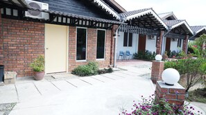 A brick building with a beige door, a covered porch with chairs, and a small garden area with plants and flowers. - Pondok Muara Chalet (Langkawi)
