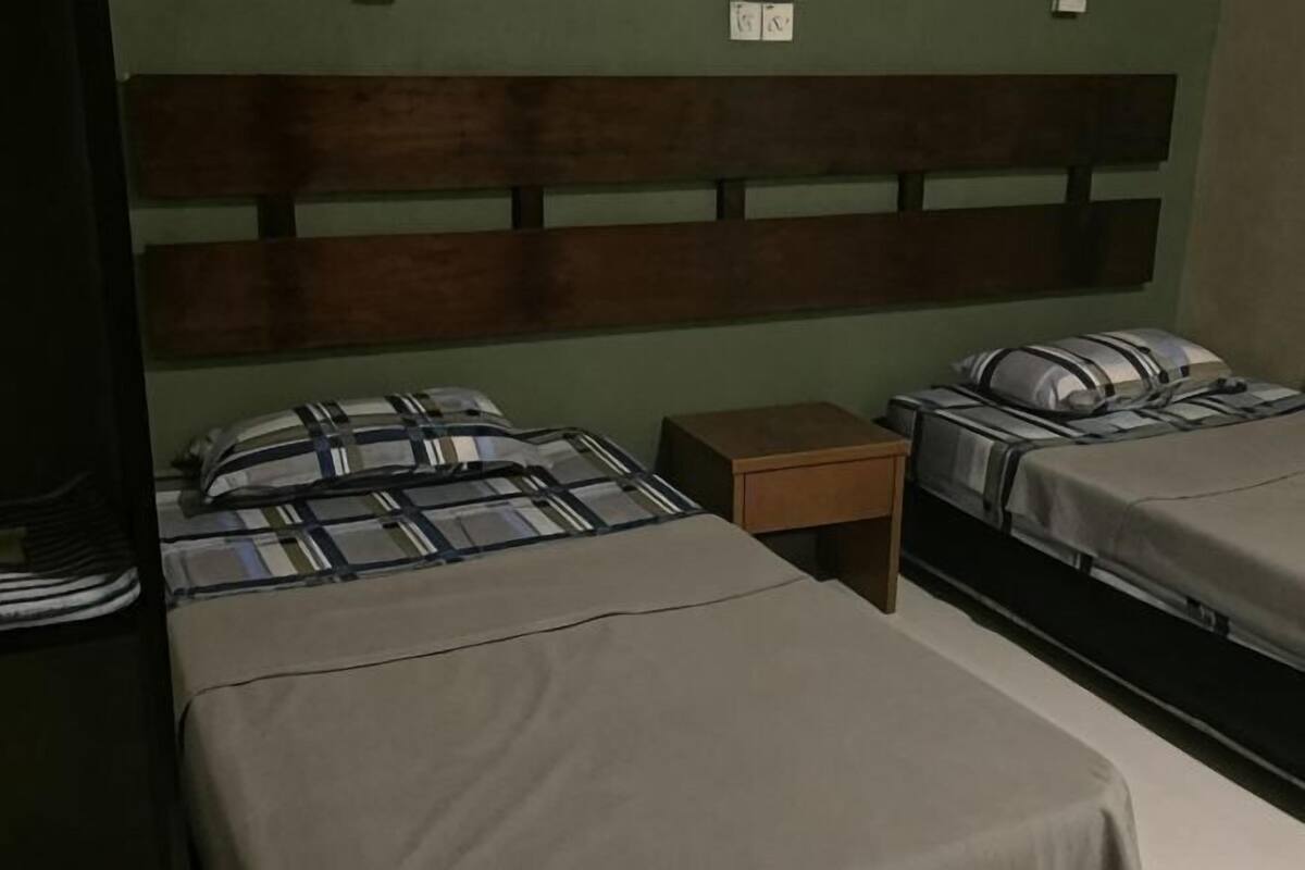 Quarto com duas camas, uma cabeceira de madeira, uma mesa de cabeceira pequena de madeira e uma tomada na parede.