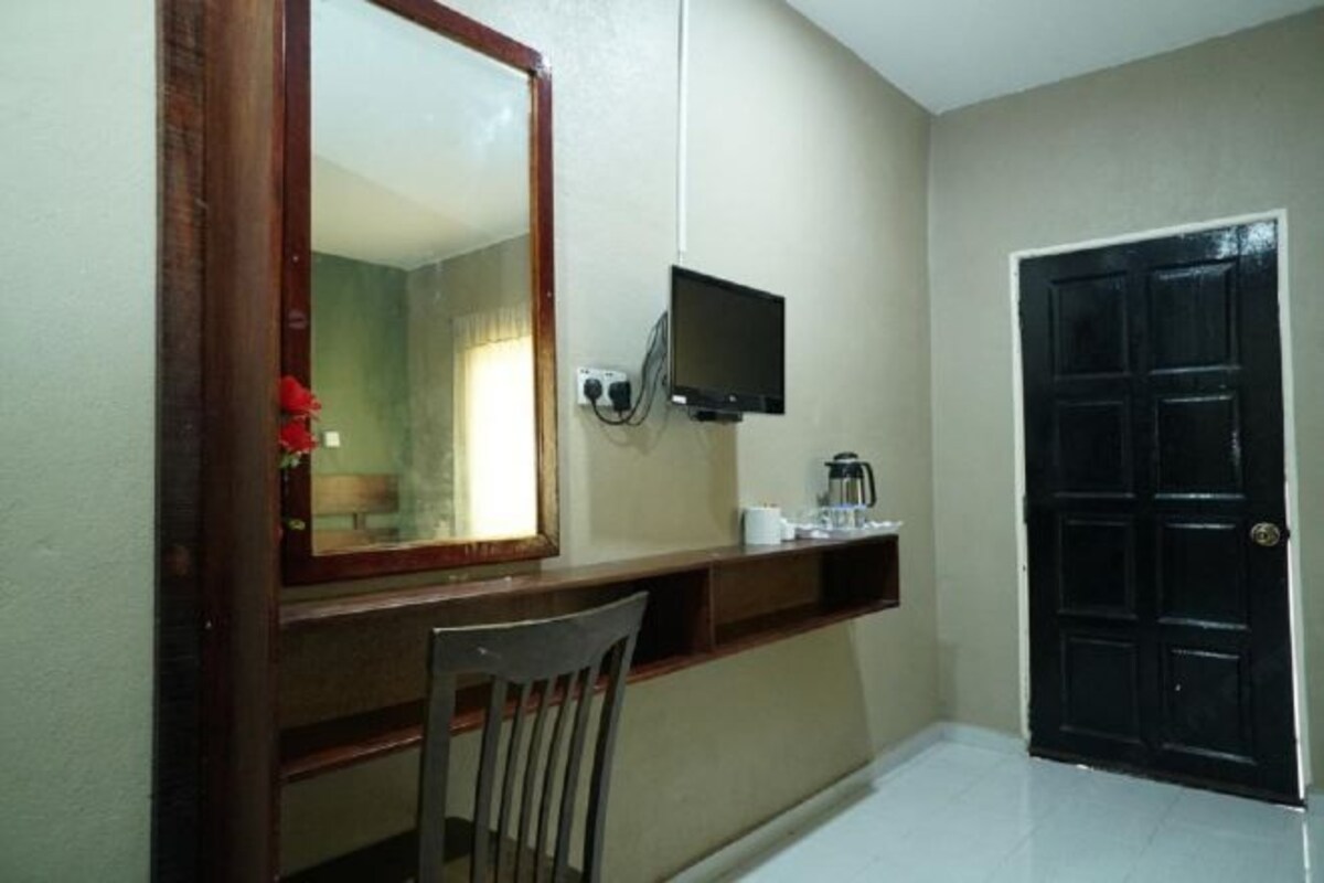 A room with a wooden desk, a chair, a television mounted on the wall, a black door, and a small shelf with items.