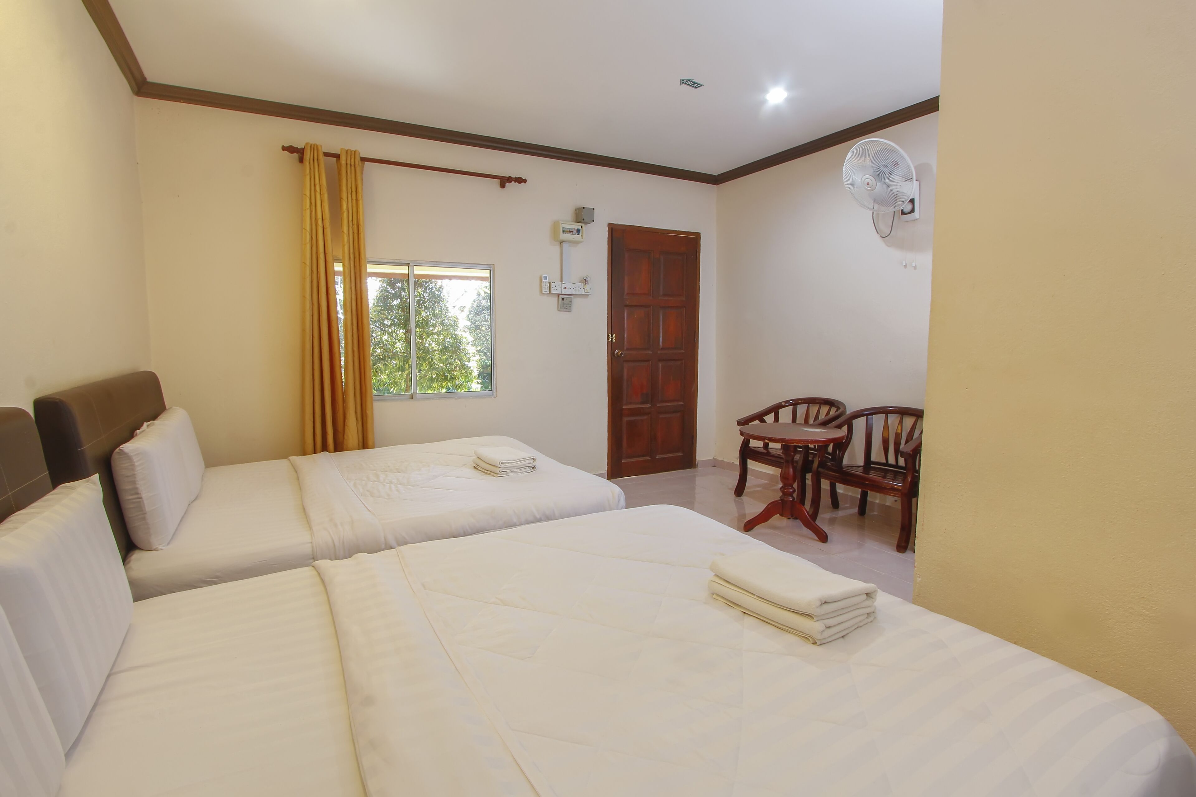 A hotel room with two beds, a wooden door, a window with curtains, a fan, and a small table with chairs.
