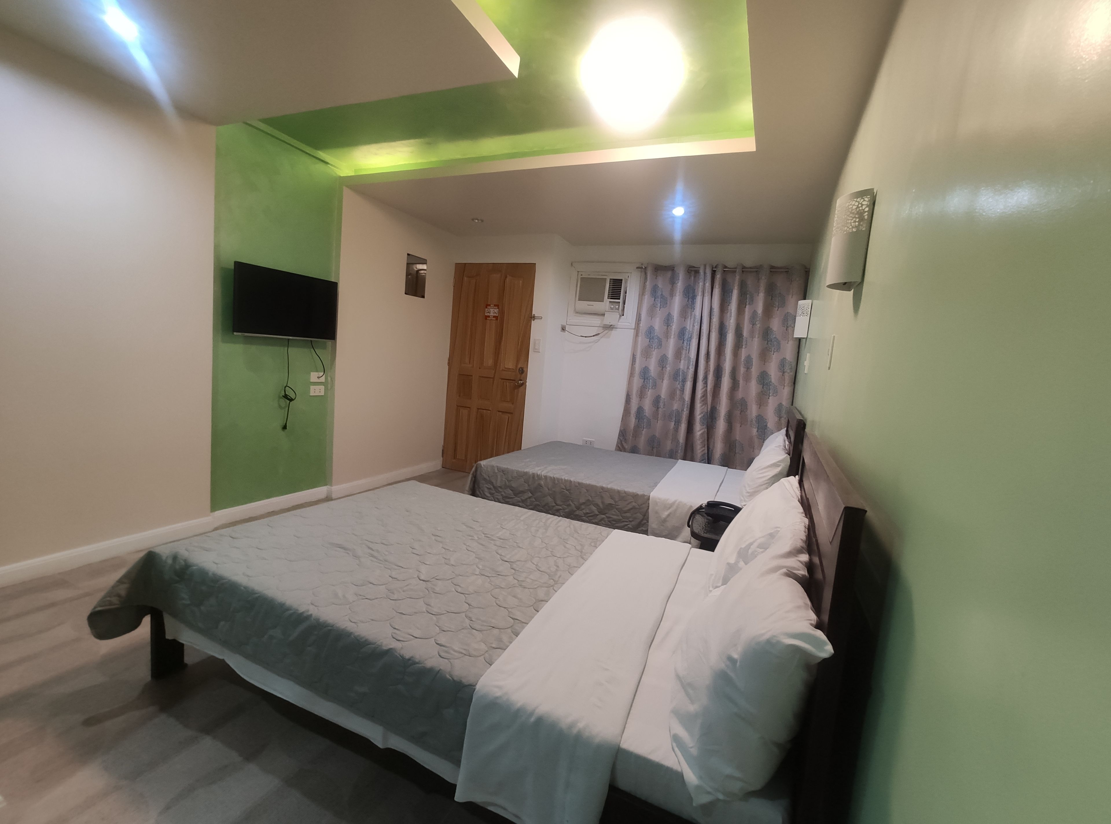 Ein Schlafzimmer mit zwei Betten, einer grünen Akzentwand, einer Deckenleuchte und einem Fenster mit Vorhängen.