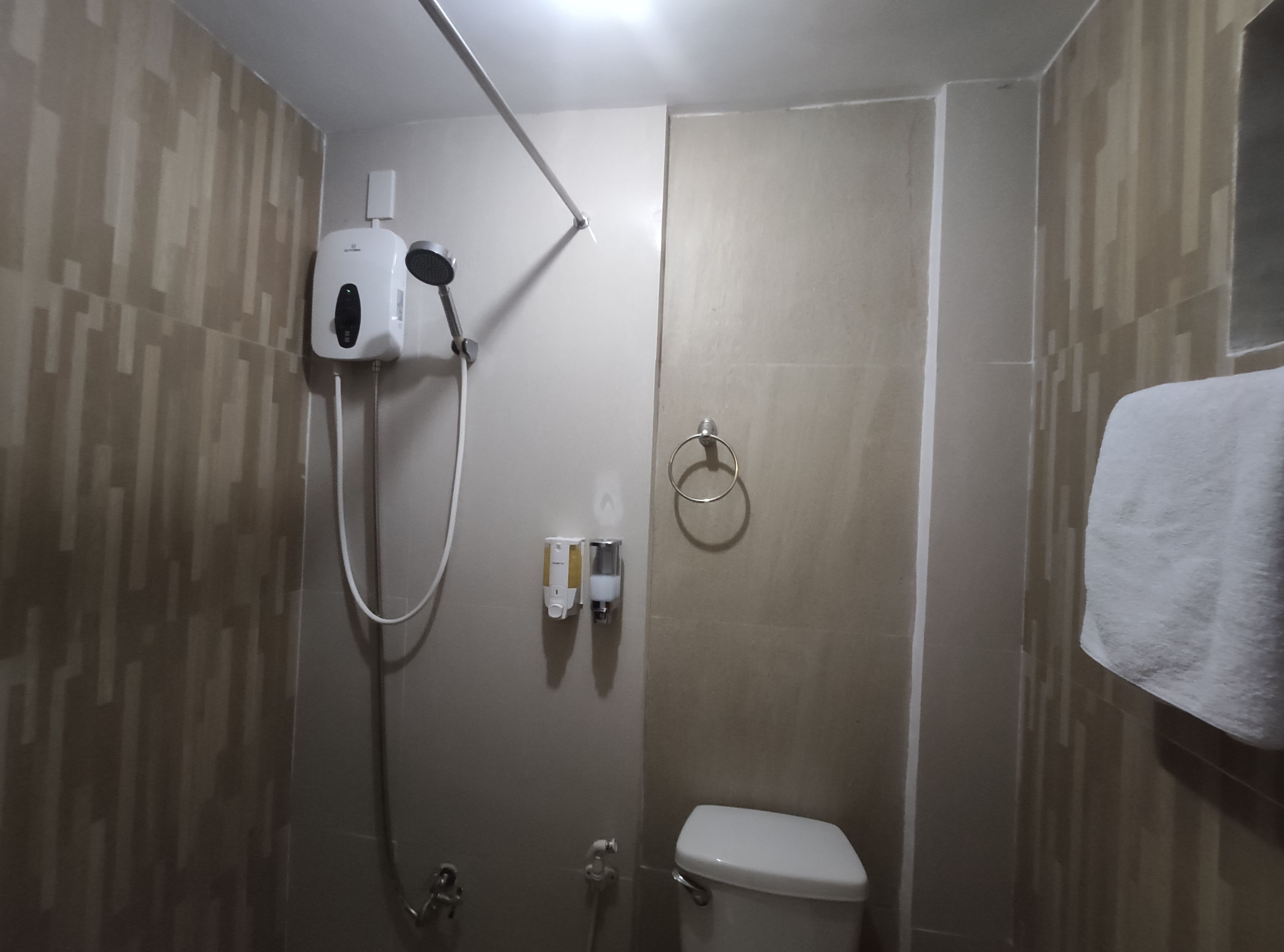A small, enclosed shower area with a toilet, a towel hanging on the wall, and a handheld showerhead mounted on the wall.