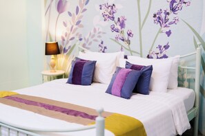 A bed with white bedding and purple pillows, a bedside table with a lamp, and a floral wall mural. - Play La Ploen Boutique Resort (Khu Mueang)