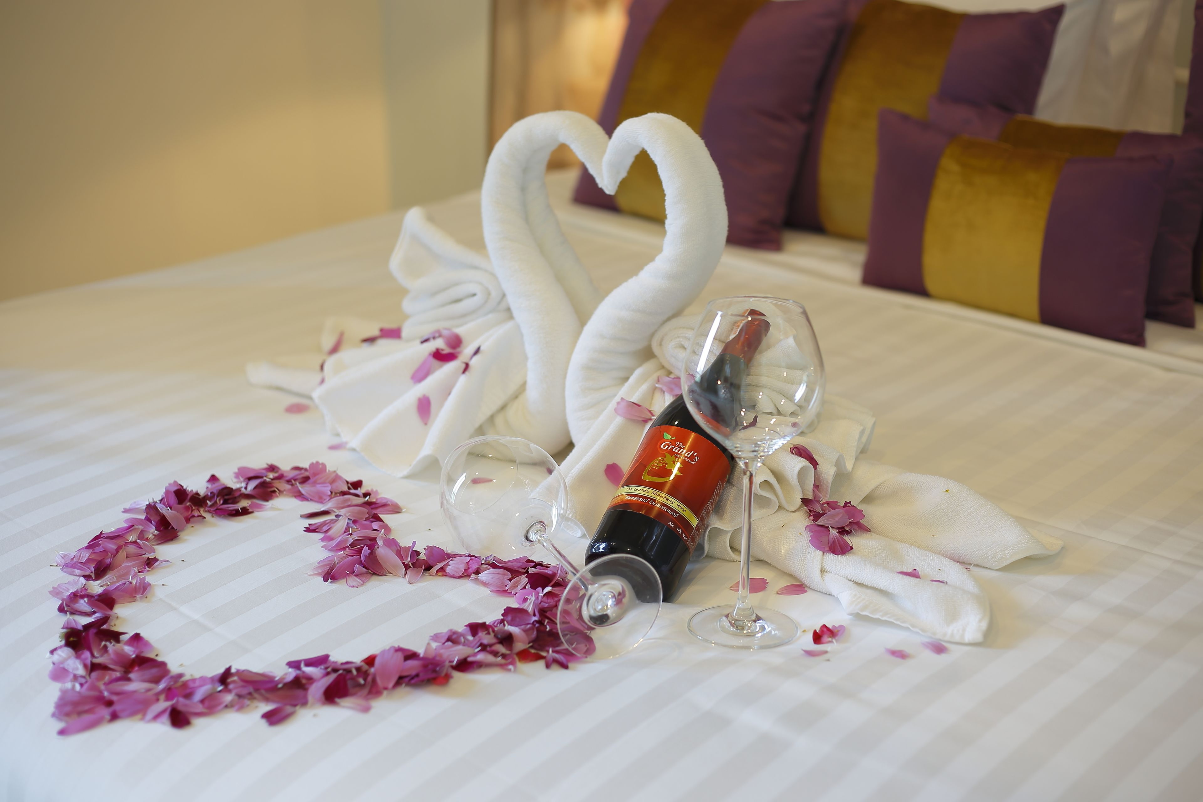 Una cama bien hecha con un arreglo de toallas en forma de corazón y pétalos de rosa, una botella de vino y dos copas de vino.