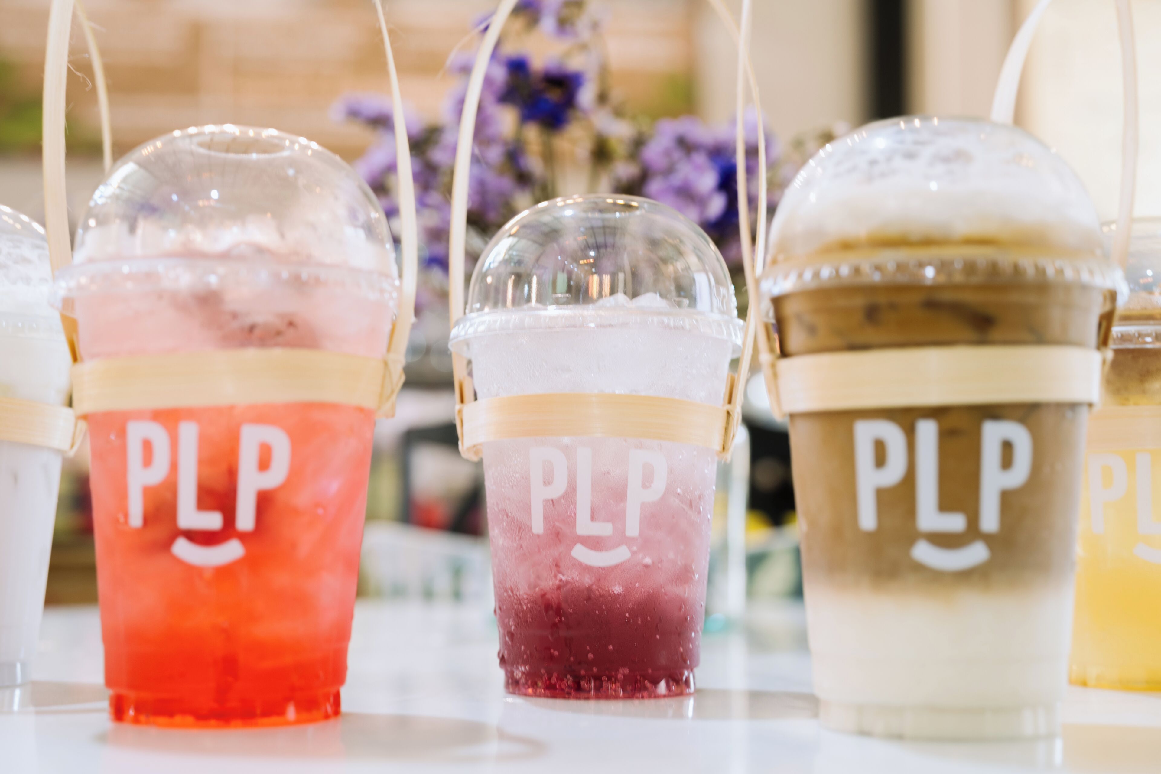 Três bebidas geladas com a marca 'PLP', cada uma em um copo de cor diferente.