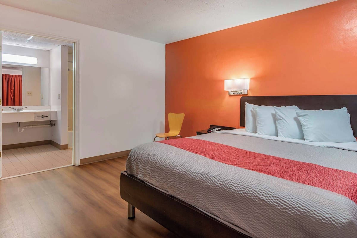 A hotel room with an orange accent wall, a bed with multiple pillows, a bedside table, a chair, and a bathroom visible through an open door.
