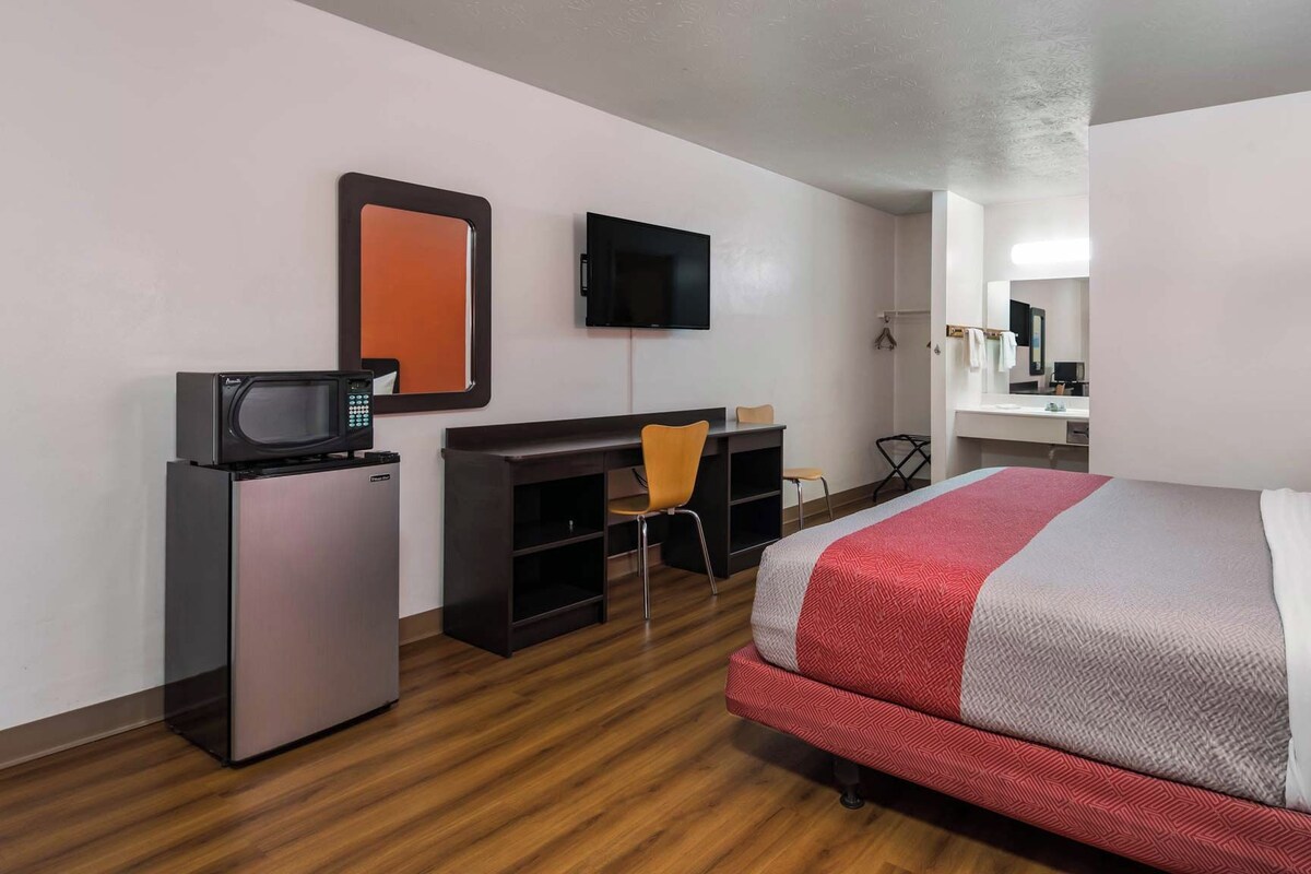 Una habitación de hotel con cama, escritorio con silla, televisión, refrigerador y espejo.