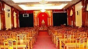 Une grande salle dotée de rangées de chaises en bois et d’un tapis rouge menant à une scène surmontée d’un décor rouge et orné d’une statue dorée.