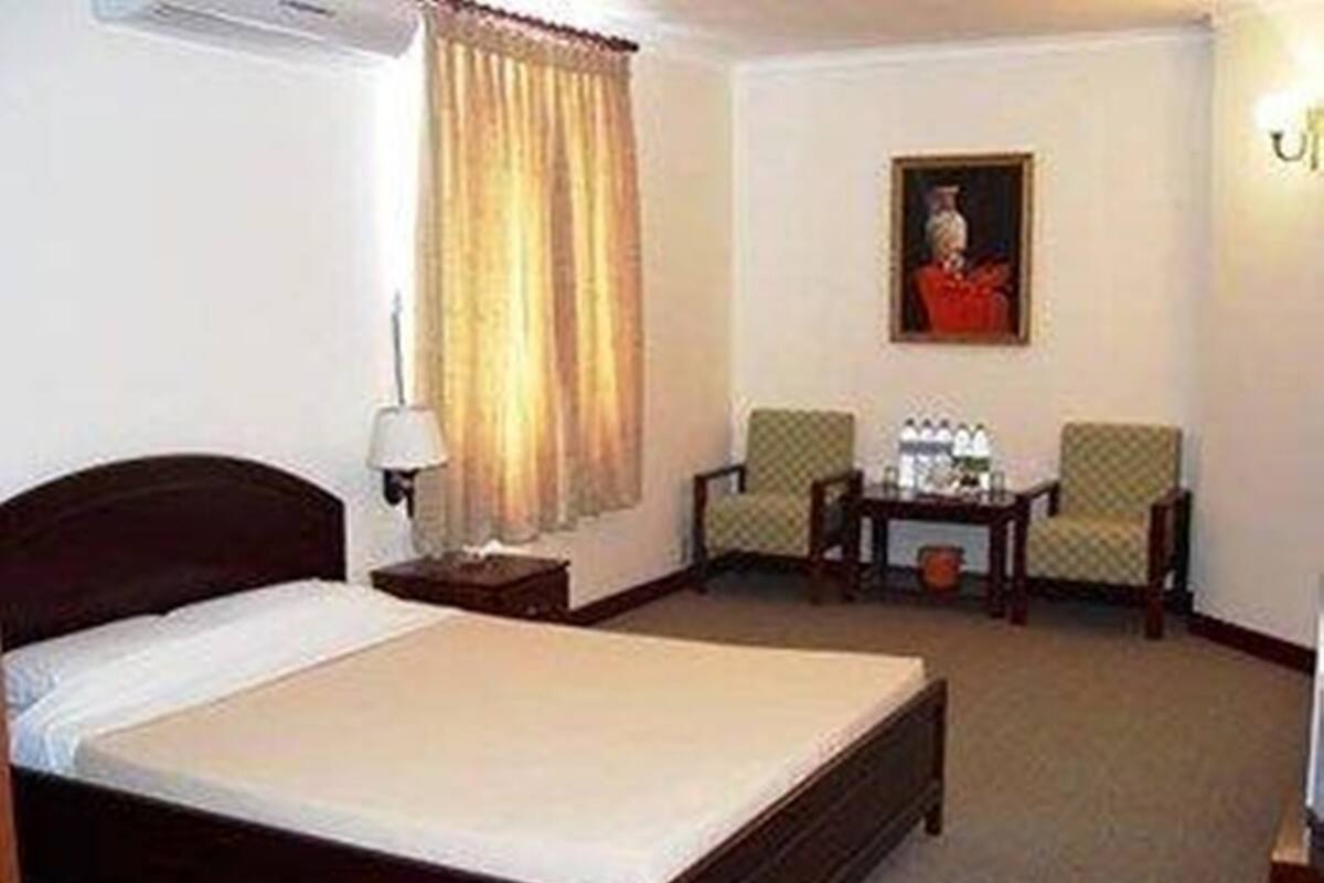 A hotel room with a bed, a nightstand, a lamp, two chairs, a small table with a bottle of water, and a painting on the wall.