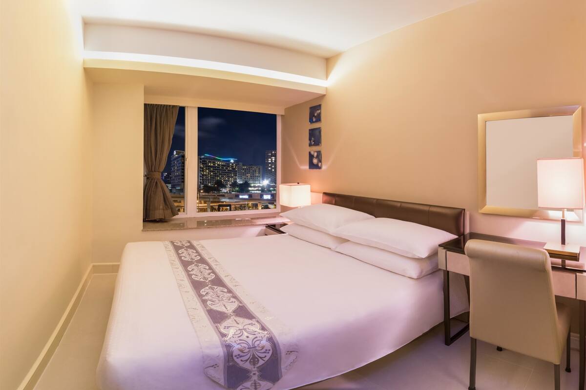 A hotel room with a large bed, a desk with a chair, a nightstand, and a view of the cityscape at night.