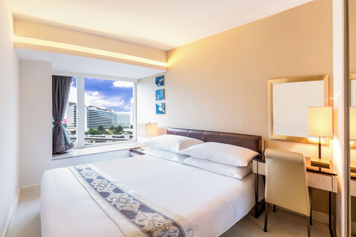 A hotel room with a large bed, a desk, a chair, a window with a view of buildings, and a wall-mounted mirror.