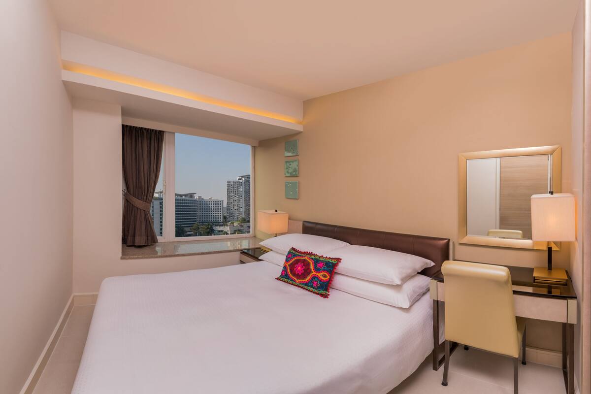 A hotel room with a bed, a desk, a chair, a window with curtains, a lamp, and a city view.