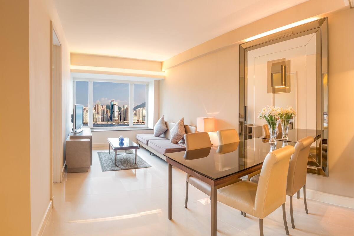 A modern living room with a glass dining table, comfortable seating, a large mirror, and a view of a cityscape through a large window.