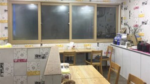 Uma zona de refeições com mesa e cadeiras de madeira, janela com persianas e bancada de cozinha com diversos objetos.