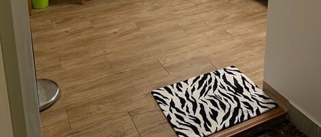 A compact bedroom with a wooden desk, a small stool, a white bedside table, a zebra-patterned rug, and a bed.