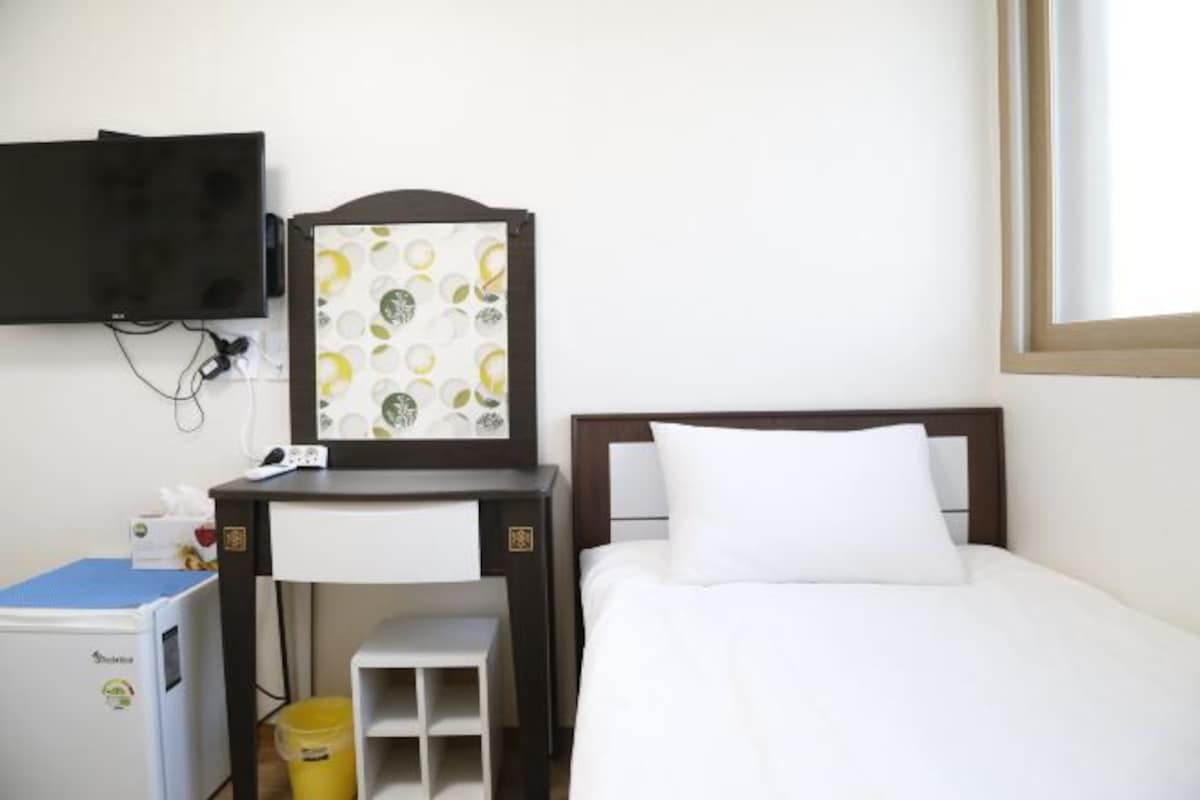 A hotel room with a bed, a desk with a chair, a television mounted on the wall, and a small trash bin.